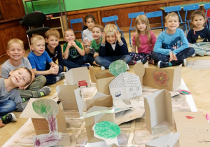 Grupa dzieci przedszkolnych siedzi na podłodze na dużej sali zabaw. Uśmiechają się prezentują swoją wspólna prace plastyczno - konstrukcyjną przedstawiającą miasto z terenami zielonymi i ciekawą architekturą. Widoczna na zdjęciu duma dzieci z wykonanego zadania.